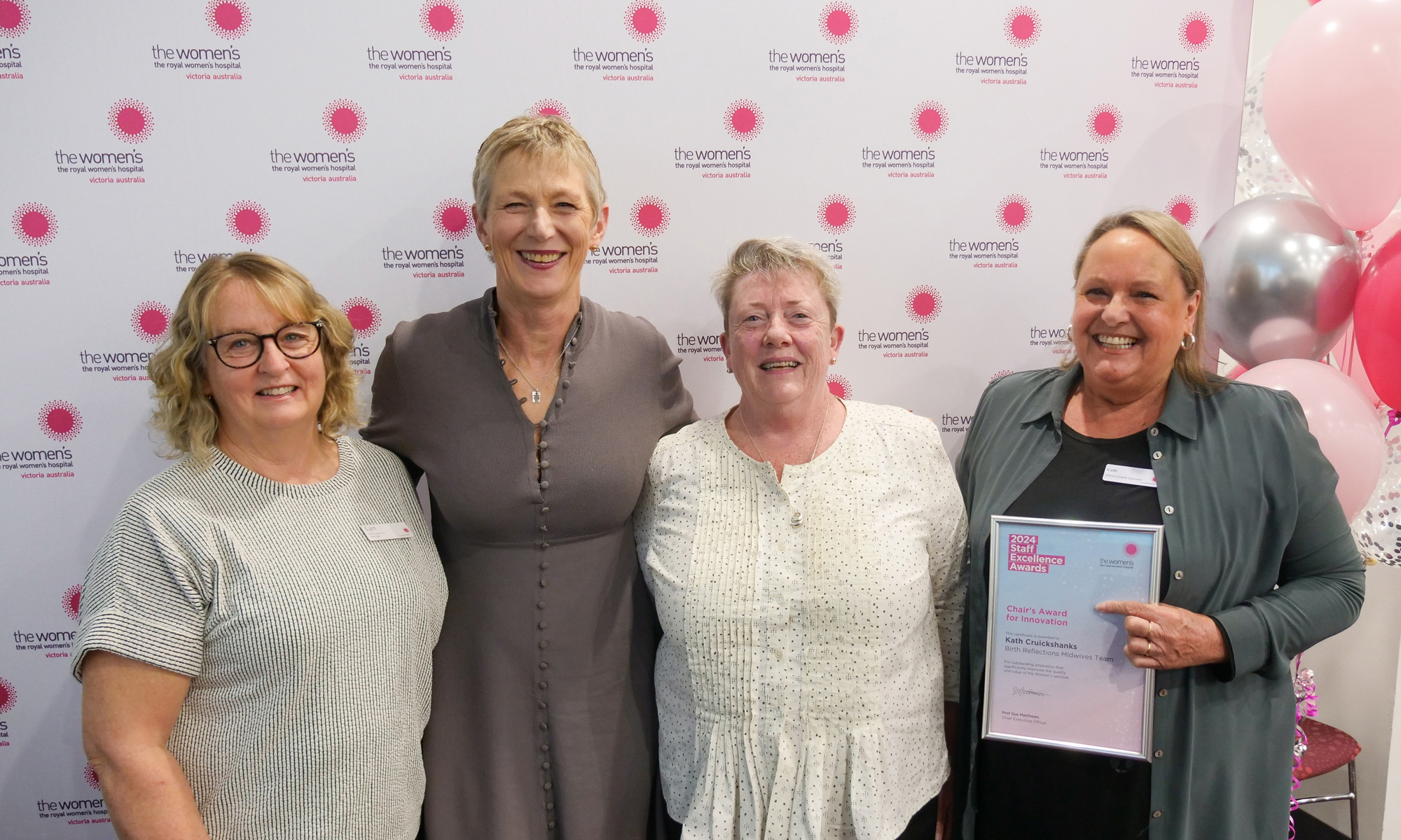 A group of four smiling people in front of the Women's signage and balloons. One is holding a Staff Excellence Awards certificate.