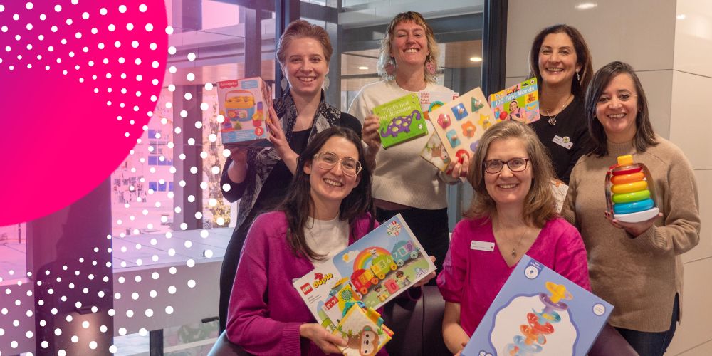 Six staff members of the First Thousand Days Clinic are holding up toys and smiling into the camera.  