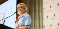 Rosie Batty standing at a lectern and speaking into the microphone, wearing a light blue dress and glasses.