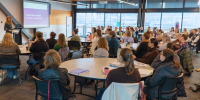  large professional workshop or seminar taking place in a modern conference room. A presenter stands at the front beside a projection screen