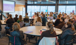  large professional workshop or seminar taking place in a modern conference room. A presenter stands at the front beside a projection screen