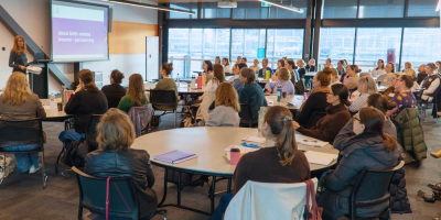  large professional workshop or seminar taking place in a modern conference room. A presenter stands at the front beside a projection screen