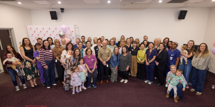 Recipients of the Women’s Length of Service award stand in a group. Some are holding kids and babies. There are balloons and a media wall wi