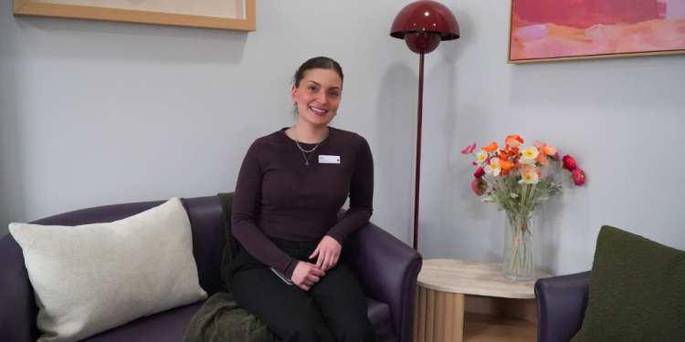 Social worker Faith sits on a couch surrounded by cushions and blankets. There is a vase with flowers in it and a lamp beside her and art on